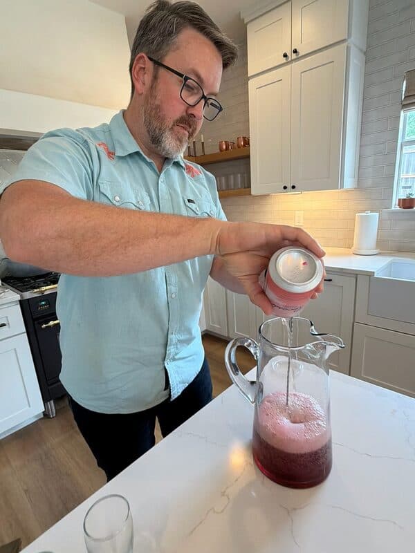 A dad pouring Recess Strawberry Rosé into a glass pitcher to mix the Strawberry Mimosa mocktail