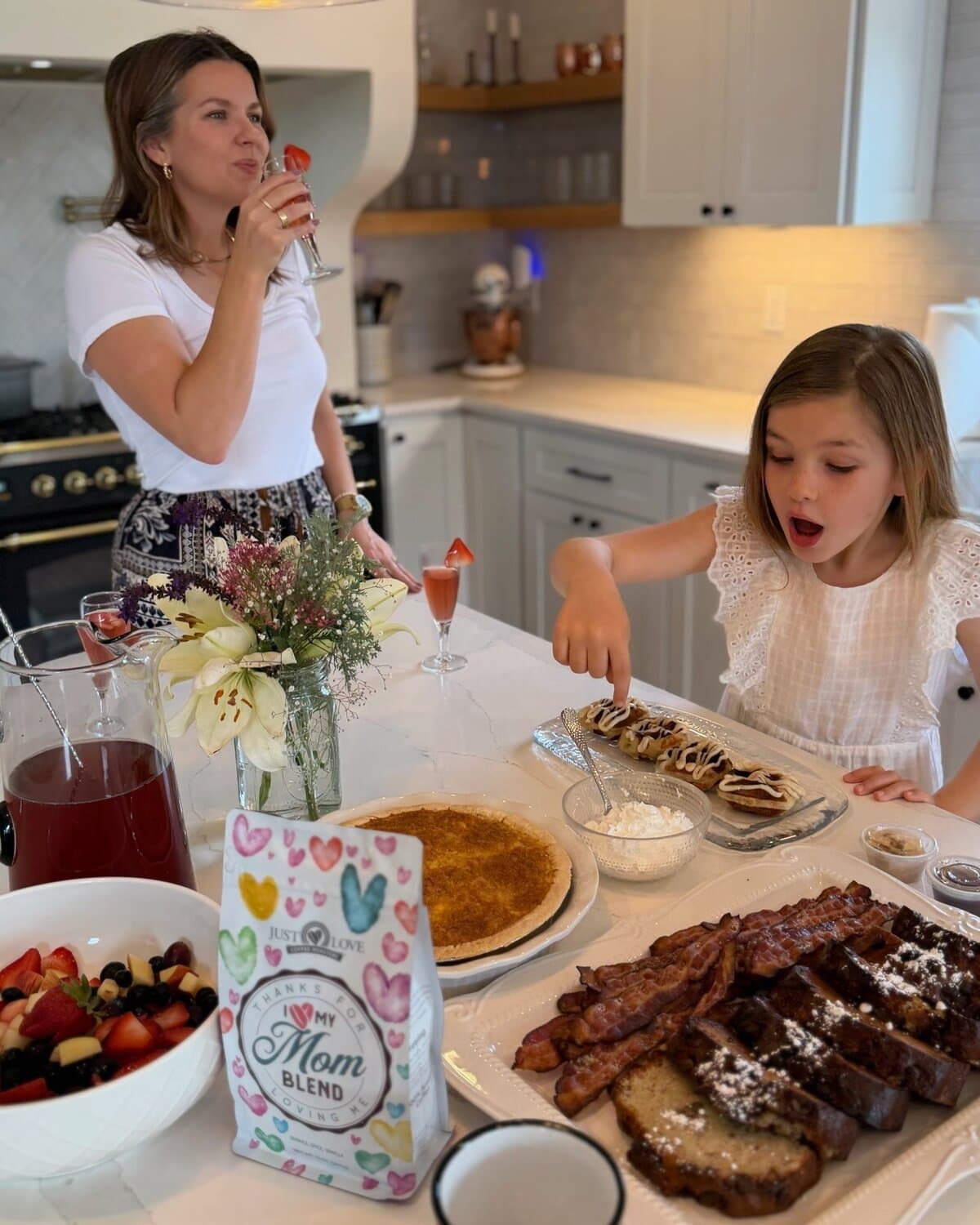 Mom sipping a Strawberry Mimosa mocktail while her daughter sneaks a cinnamon roll from the 2026 Brunch Kit spread